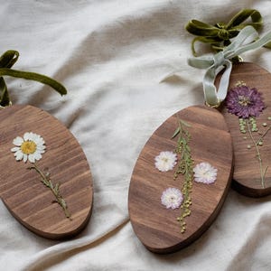May include: Three wooden oval ornaments with dried flowers. One features a white daisy, another has small white flowers and greenery, and the third displays a purple flower. Each ornament has a green velvet ribbon for hanging.