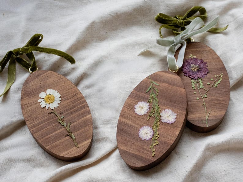 May include: Three wooden oval ornaments with dried flowers. One features a white daisy, another has small white flowers and greenery, and the third displays a purple flower. Each ornament has a green velvet ribbon for hanging.