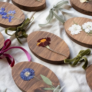 May include: Oval wooden ornaments with pressed flowers in various colors, including blue, white, and purple. Each ornament is adorned with a velvet ribbon in shades of green, pink, and purple. The ornaments are arranged on a white fabric surface.