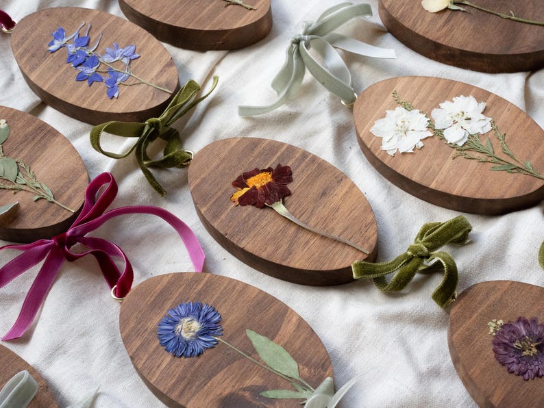 May include: Oval wooden ornaments with pressed flowers in various colors, including blue, white, and purple. Each ornament is adorned with a velvet ribbon in shades of green, pink, and purple. The ornaments are arranged on a white fabric surface.