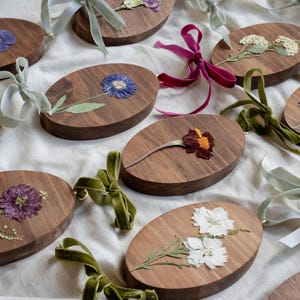 May include: Oval wooden ornaments with pressed flowers in various colors, including blue, purple, and white. Each ornament has a velvet ribbon for hanging. The ornaments are arranged on a white surface.