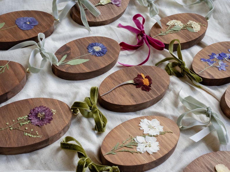 May include: Oval wooden ornaments with pressed flowers in various colors, including blue, purple, and white. Each ornament has a velvet ribbon for hanging. The ornaments are arranged on a white surface.