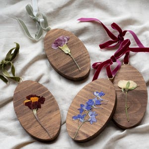 May include: Oval wooden ornaments with dried flowers, each with a velvet ribbon for hanging. The flowers include a marigold, blue delphiniums, a carnation, and a rose. The ribbons are in shades of green, burgundy, and light grey.