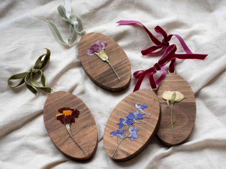 May include: Oval wooden ornaments with dried flowers, each with a velvet ribbon for hanging. The flowers include a marigold, blue delphiniums, a carnation, and a rose. The ribbons are in shades of green, burgundy, and light grey.