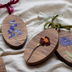 May include: Wooden oval ornaments with pressed flowers and velvet ribbon hangers. One features blue larkspur, another a marigold, and a third a violet. Text on the ornaments reads "larkspur (july)", "marigold (october)", and "violet (february)".