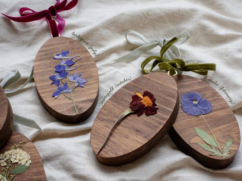 May include: Wooden oval ornaments with pressed flowers and velvet ribbon hangers. One features blue larkspur, another a marigold, and a third a violet. Text on the ornaments reads "larkspur (july)", "marigold (october)", and "violet (february)".