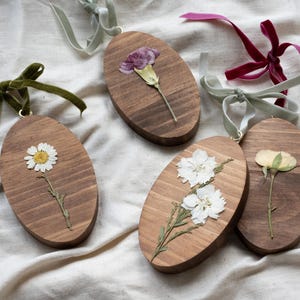 May include: Oval wooden ornaments with pressed flowers. Each ornament features a different flower, including a daisy, carnation, and rose. The ornaments are adorned with velvet ribbons in various colors, such as green, pink, and burgundy.