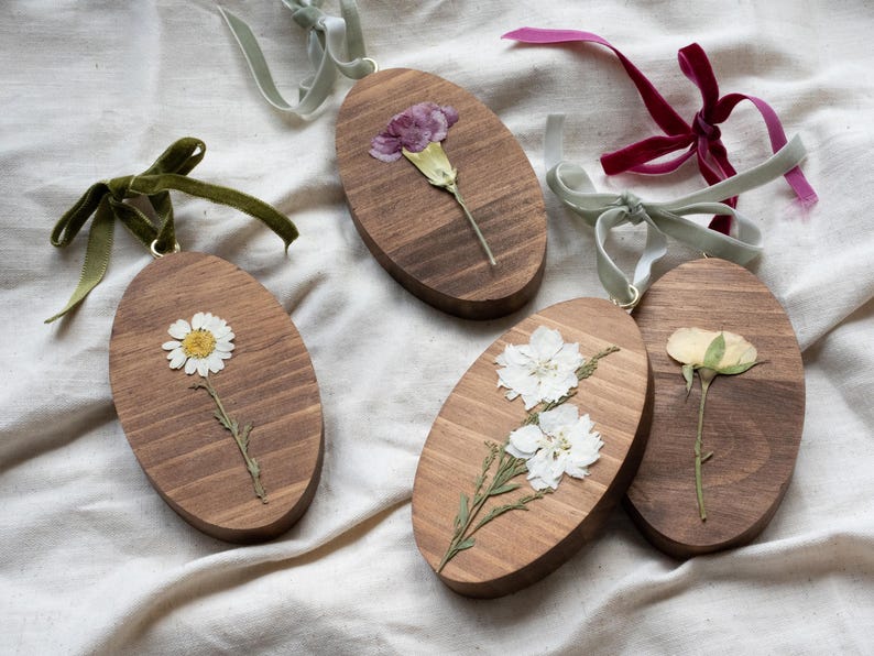 May include: Oval wooden ornaments with pressed flowers. Each ornament features a different flower, including a daisy, carnation, and rose. The ornaments are adorned with velvet ribbons in various colors, such as green, pink, and burgundy.