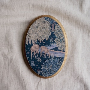 May include: A round, framed tapestry with a blue background featuring two deer in a woodland setting. The tapestry has a gold frame and a detailed design of trees, leaves, and a stream.