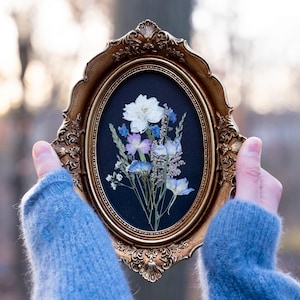 May include: An oval, ornate gold frame holds a pressed flower arrangement against a black background. The arrangement features white, blue, and purple flowers. The frame is held by hands wearing a blue sweater.