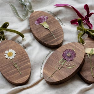 May include: Oval wooden ornaments with dried flowers, including a daisy, carnation, chrysanthemum, and rose. Each ornament has a velvet ribbon for hanging. The flowers are in shades of white, purple, and yellow.