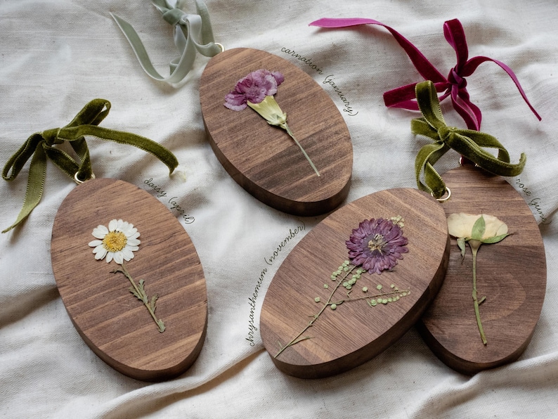 May include: Oval wooden ornaments with dried flowers, including a daisy, carnation, chrysanthemum, and rose. Each ornament has a velvet ribbon for hanging. The flowers are in shades of white, purple, and yellow.