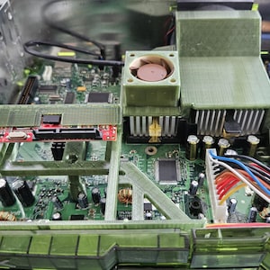 May include: The interior of an open game console featuring a green circuit board, a small cooling fan, silver heat sinks, and a custom-fitted translucent green 3D-printed plastic frame. Multicolored wiring bundles connect to the power supply unit.