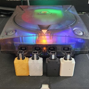 May include: A translucent black video game console with a circular disc lid and a glowing multicolored interior light. Four rectangular controller port adapters in yellow, clear, black, and white are positioned in front of the console unit.