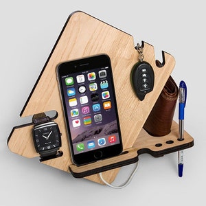 Pode incluir: Organizador de mesa de madeira com acabamento em grão de madeira clara. Ele contém um smartphone preto, um relógio preto, uma carteira marrom, uma caneta azul e um chaveiro de carro. Um cabo de carregamento branco está conectado ao telefone.
