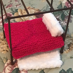 May include: A miniature bed with a red knitted blanket and a fluffy white pillow. The bed frame is made of dark metal. The bed is designed for a doll or small pet.