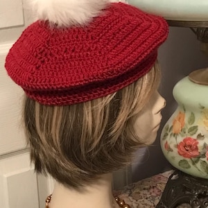 May include: A deep red crocheted beret featuring a textured, circular stitch pattern and a large, fluffy white pom-pom attached to the center top. The hat is constructed from yarn and sits atop a mannequin head, showcasing its rounded shape.