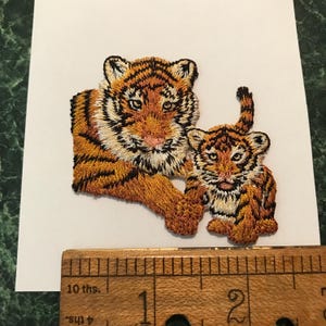 May include: An embroidered patch featuring an adult tiger and a tiger cub with orange, black, and white thread. The patch is positioned above a wooden ruler showing a width of approximately 2.5 inches for the tiger design on the white paper.