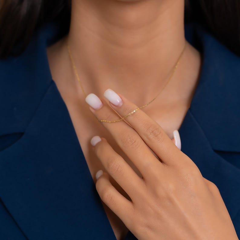 Pode incluir: Delicado colar de ouro com uma corrente fina, mostrado numa m&atilde;o. O colar &eacute; exibido contra um blazer azul escuro, criando um contraste. A corrente tem aproximadamente 45 cm de comprimento.
