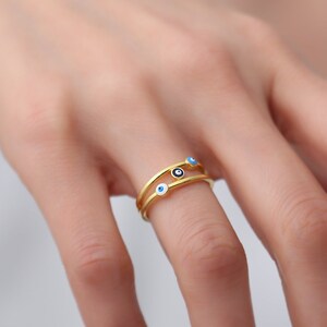 May include: Gold ring with three blue evil eye charms. The ring is worn on a finger.