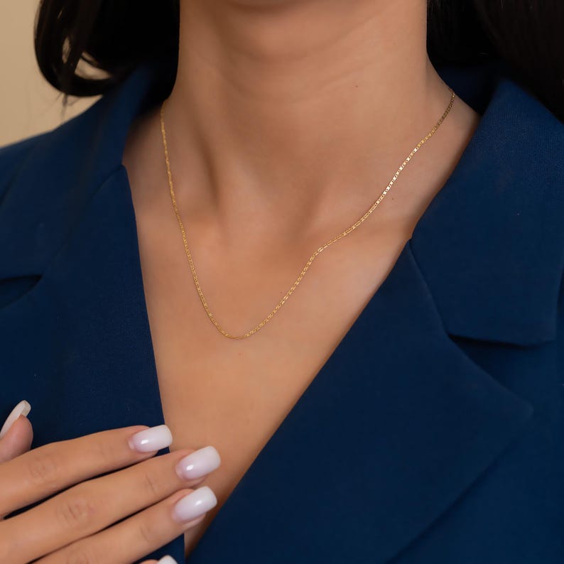 Pode incluir: Um delicado colar de ouro com uma corrente de elos retangulares. O colar &eacute; exibido contra um blazer azul escuro. A imagem mostra um close-up do colar em volta do pesco&ccedil;o.