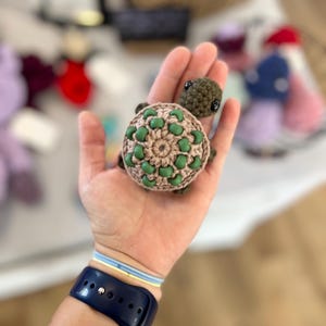 May include: A handmade crochet turtle with a tan shell adorned with green beads. The turtle's head and limbs are a dark green color. The turtle is a small, decorative item.