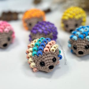 May include: A collection of crocheted hedgehogs with colorful beaded spines. The hedgehogs are in various colors, including purple, orange, yellow, blue, and pink. The beads are arranged in a spiral pattern on the spines.