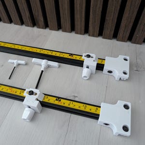 Puede incluir: Dos rieles de medición negros con cintas métricas amarillas, soportes de montaje blancos y herramientas en forma de T. Las cintas métricas muestran marcas en pulgadas. El fondo es un panel de madera.