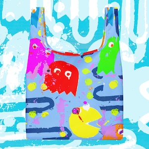 May include: A reusable shopping bag with a light blue background featuring colorful cartoon ghosts and Pac-Man characters. The ghosts are in pink, red, and green, with yellow dots and blue lines scattered across the bag. The bag has a red trim.