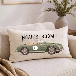 May include: A rectangular throw pillow with a beige textured fabric cover. The pillow features a printed illustration of a vintage sage green convertible sports car with the number 11 on the side and the black text Noah's Room above it.