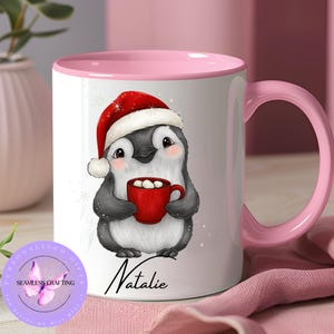 May include: White ceramic mug with a pink interior and handle. The mug features a cartoon penguin wearing a Santa hat and holding a red mug of hot chocolate. The name "Natalie" is written in cursive below the penguin.