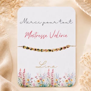 Peut inclure: Une carte blanche avec le texte "Merci pour tout Maîtresse Valérie Lina" en script noir et rose. Un bracelet de perles avec des accents dorés est attaché à la carte. Le bas de la carte présente un motif floral avec des fleurs roses, bleues et jaunes.