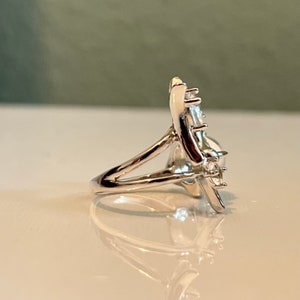 May include: A silver ring with a floral design, featuring a large, clear gemstone at the centre and smaller stones around it. The ring's band is split, creating an open design. The ring is displayed on a white surface.