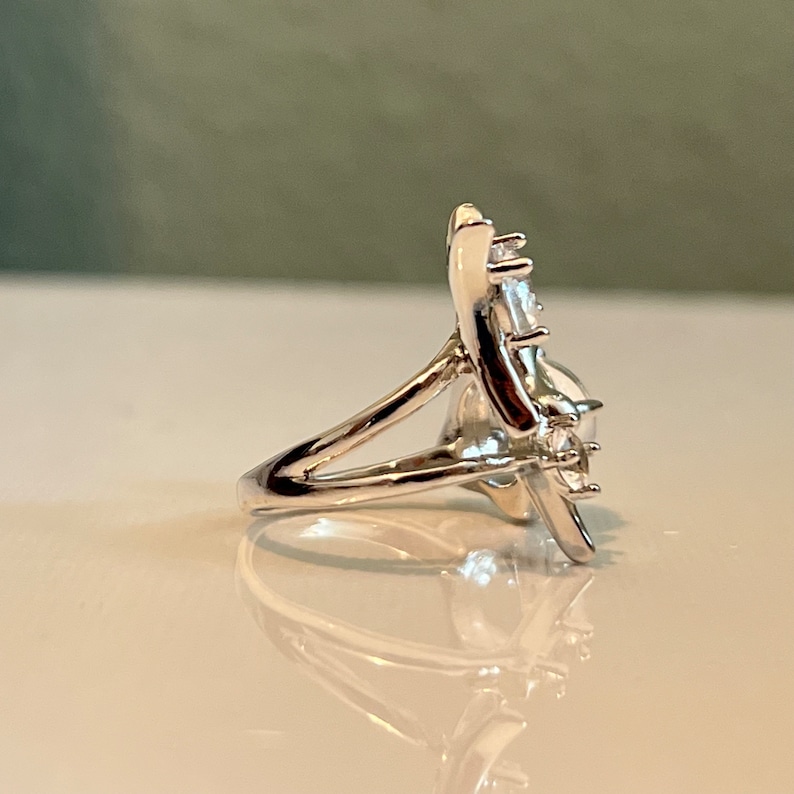 May include: A silver ring with a floral design, featuring a large, clear gemstone at the centre and smaller stones around it. The ring's band is split, creating an open design. The ring is displayed on a white surface.