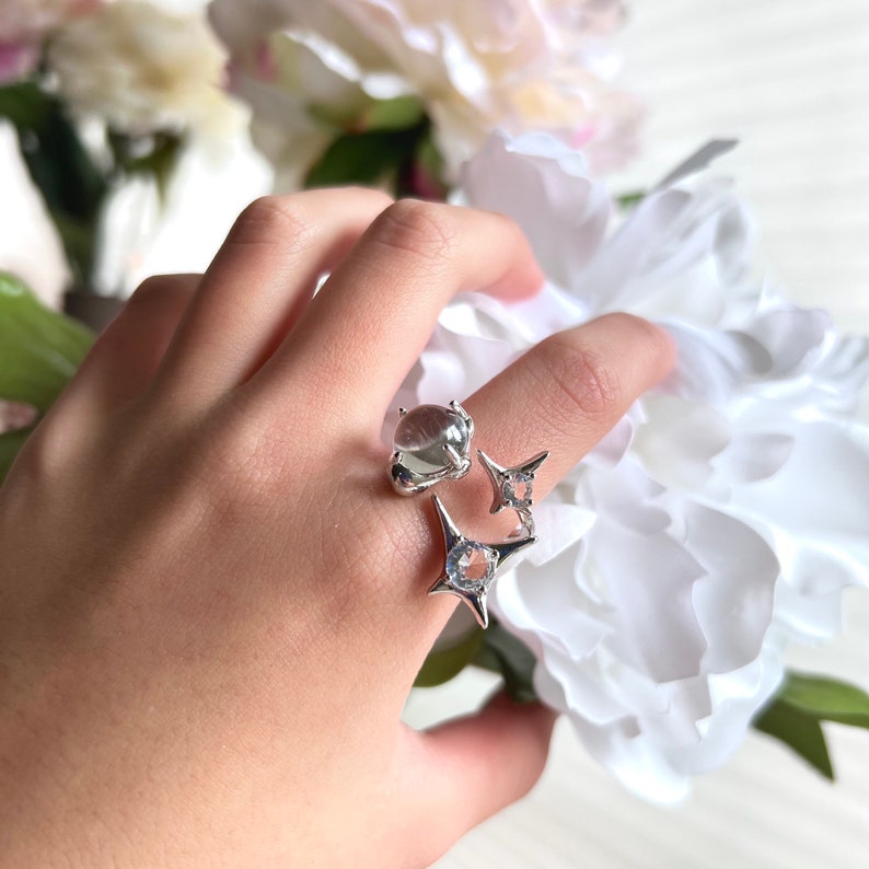 May include: A silver ring with three distinct designs. One features a clear, rounded stone, while the others showcase starburst shapes with clear, sparkling stones. The ring is displayed on a finger, with white flowers in the background.