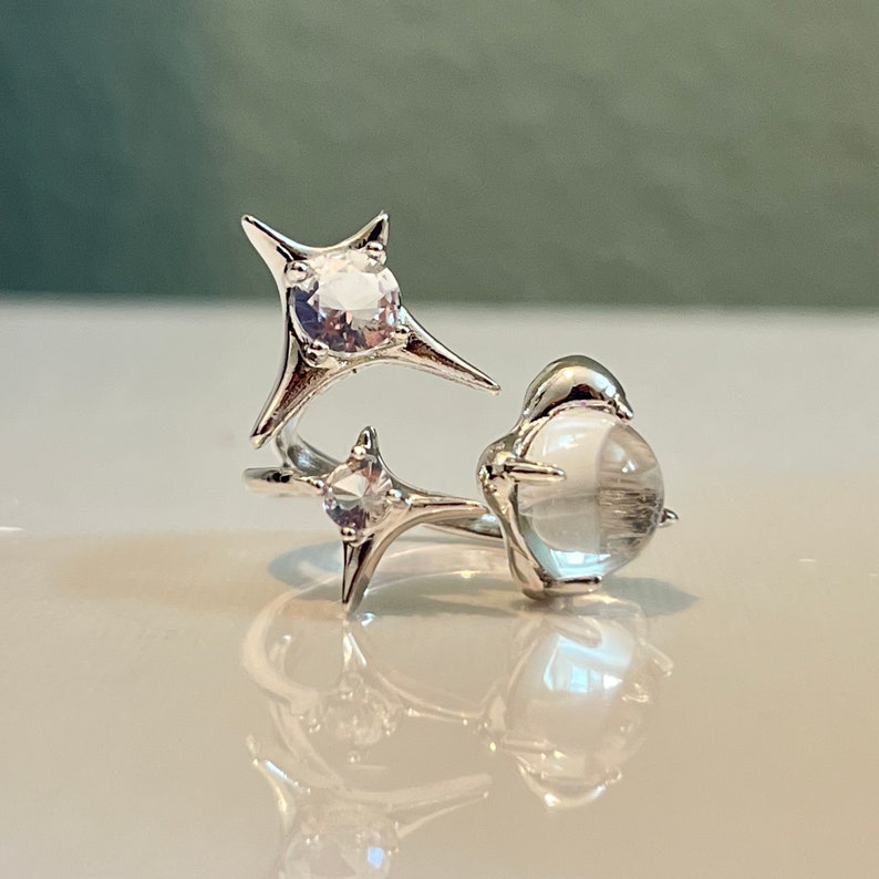 May include: A silver ring with a distinctive design. It features three clear, round gemstones and two star-shaped accents. The gemstones are set in a delicate, openwork setting, creating a modern and elegant appearance.