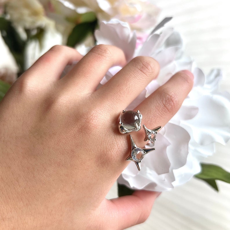 May include: A silver ring featuring a square, grey-toned gemstone and two star-shaped embellishments with clear stones. The ring is displayed on a finger, with white flowers in the background.
