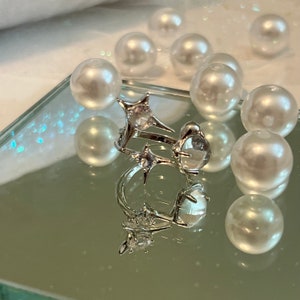 May include: Two silver rings with star and clear stone details are reflected on a mirrored surface. White pearl beads surround the rings, creating a delicate and elegant aesthetic. The rings are likely fashion jewellery.
