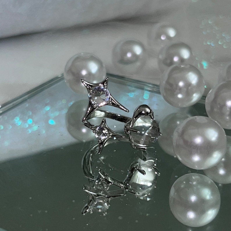 May include: A silver ring with a starburst design and clear gemstones. The ring features a large, central gemstone and smaller accent stones. The ring is displayed on a reflective surface with white pearls in the background.
