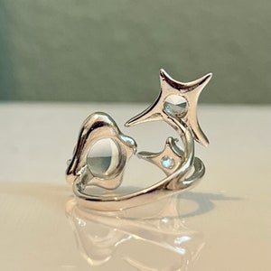 May include: A silver ring with an abstract design. The ring features a rounded shape and a starburst design with a small, clear stone in the centre of each shape. The ring is on a reflective surface.