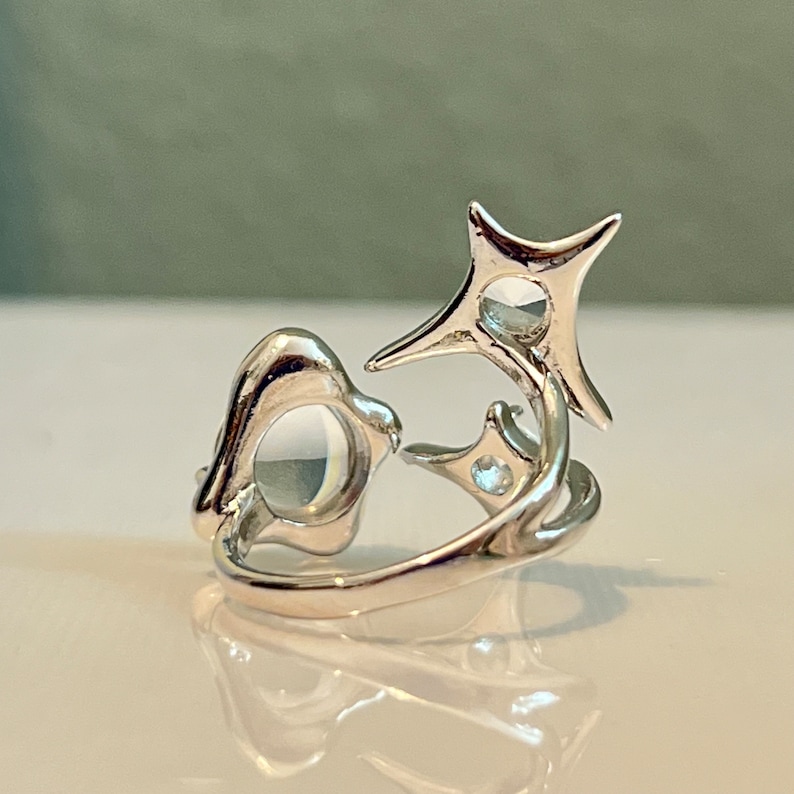 May include: A silver ring with an abstract design. The ring features a rounded shape and a starburst design with a small, clear stone in the centre of each shape. The ring is on a reflective surface.