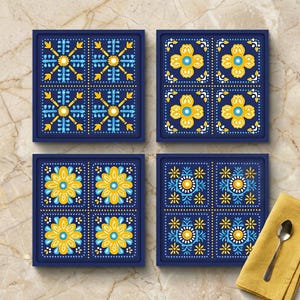 May include: A set of four square coasters with a dark blue background and intricate yellow, light blue, and white floral and geometric patterns. Each coaster features a grid of four distinct designs, characterized by symmetrical motifs and dots.
