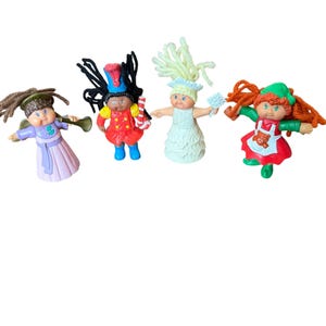 May include: Four small plastic dolls with yarn hair. One wears a purple dress and holds a horn, one is a soldier in a red uniform with a candy cane, one wears a white dress with a snowflake, and one wears a red dress with a white apron.