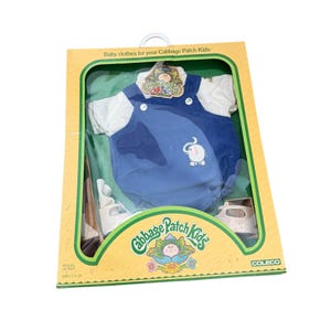 May include: A Cabbage Patch Kids baby clothes set in original packaging, featuring a white short-sleeved shirt and blue corduroy overalls with an elephant graphic. The yellow box includes the Cabbage Patch Kids logo and Coleco branding.