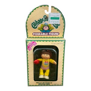 May include: A Cabbage Patch Kids poseable figure in original packaging. The doll has brown hair, a yellow long-sleeve shirt with a diamond pattern, and pink pants. The box features the text Cabbage Patch Kids, Poseable Figure, First Edition, and Hello, My Name Is Melissa Ann.