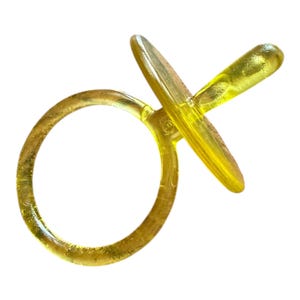 May include: A translucent yellow plastic pacifier featuring a circular handle attached to a flat, round shield and a protruding nipple. The material has a slightly textured, semi-clear appearance, allowing light to pass through the yellow surface.