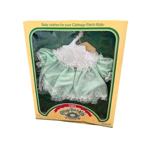 May include: A mint green doll dress with white lace trim on the collar and sleeves, displayed on a white plastic hanger inside a yellow cardboard box labeled Baby clothes for your Cabbage Patch Kids and 1984 Cabbage Patch Kids.