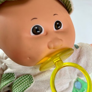 May include: A Cabbage Patch Kid doll wearing a white and green checkered hat and a white outfit with green checkered accents. The doll is holding a yellow pacifier.
