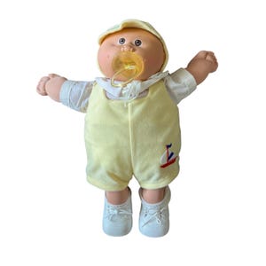 May include: A doll with a round face, tan skin, and yarn hair wearing a yellow cap, a white shirt, and yellow overalls featuring a small sailboat graphic. The doll has a translucent yellow pacifier in its mouth and wears white shoes.