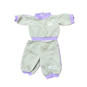 May include: A two-piece outfit consisting of a light green long-sleeve pullover top and matching pants. The top features a lavender mock neck, lavender cuffs, and a small white cat graphic. The pants have lavender cuffs and a small rectangular label.